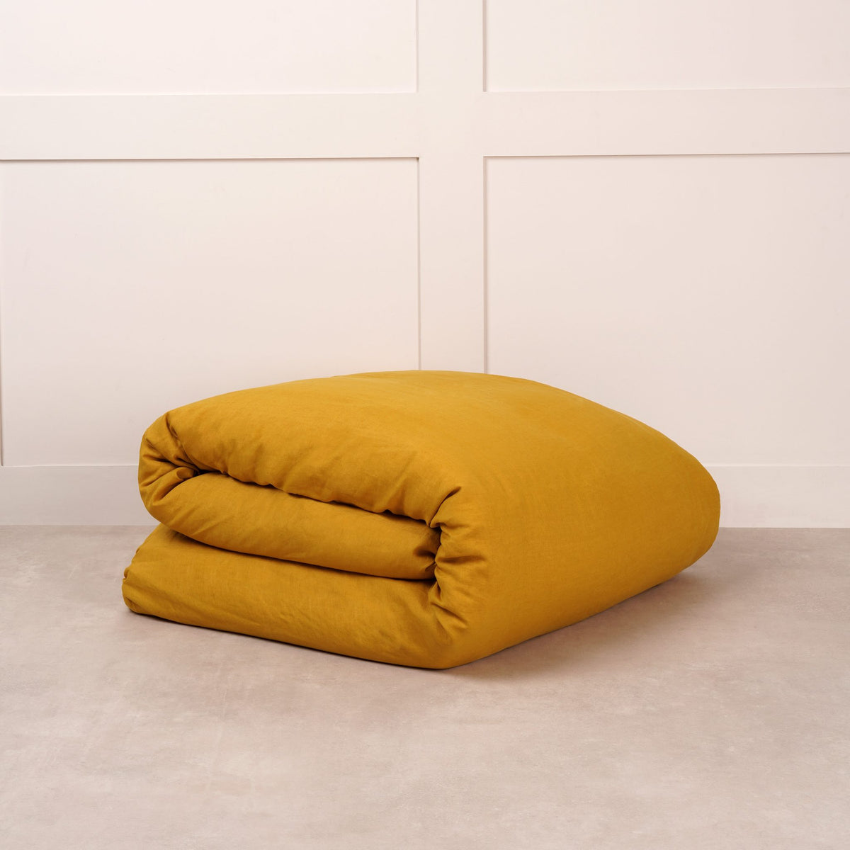 Cuddledown linen duvet cover in Ochre, neatly folded on a stone floor against a white panelled wall